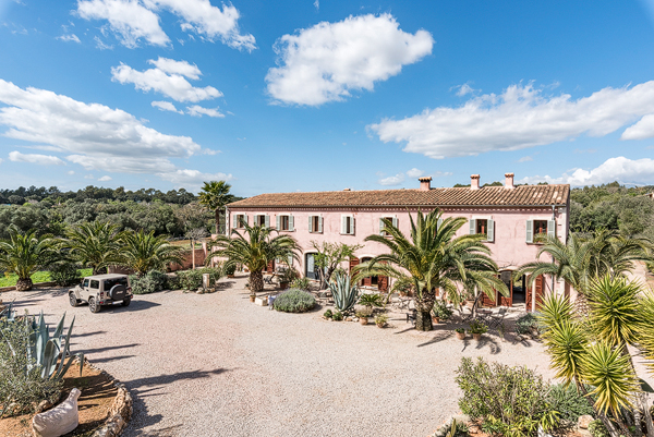 The long pink facade of Finca Can Pina seen across its gravel courtyard, with palms and a low stone wall under a wide Mallorcan sky