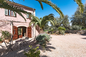 A corner of the pink finca with green shutters, an outdoor table for two and a tall palm leaning over the gravel