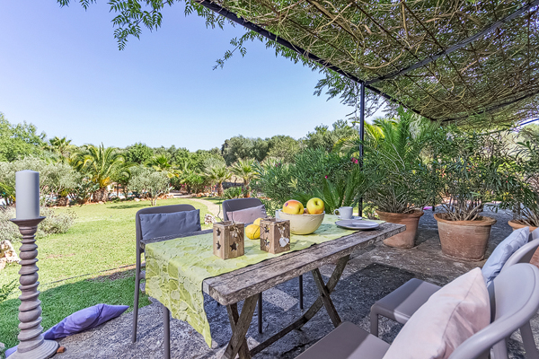 A weathered wooden table laid under a reed canopy at the edge of the garden, with a bowl of apples, lanterns and a candle, looking out over the lawn and palms