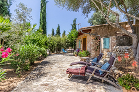 A stone cottage at the finca with green shutters, a paved terrace, two reclining chairs and beds of bougainvillea framed by tall cypress and olive