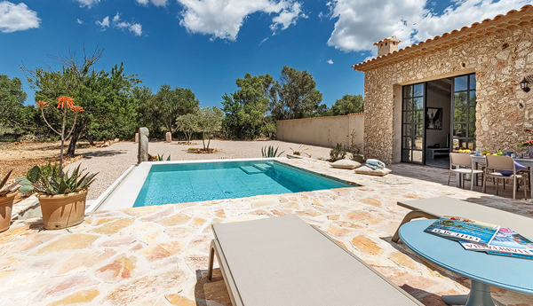 The finca's pool with a fan of golden stone paving, sun-loungers, and a stone outbuilding with glass doors opening to a dining table